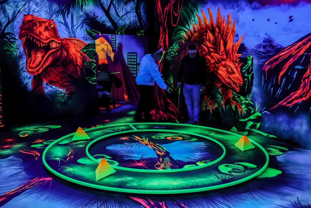 Eine fantasievoll gestaltete Minigolfbahn zeigt ein grünes Hindernis mit Looping unter buntem UV-Licht, umgeben von knallig bemalten Pflanzenwänden.