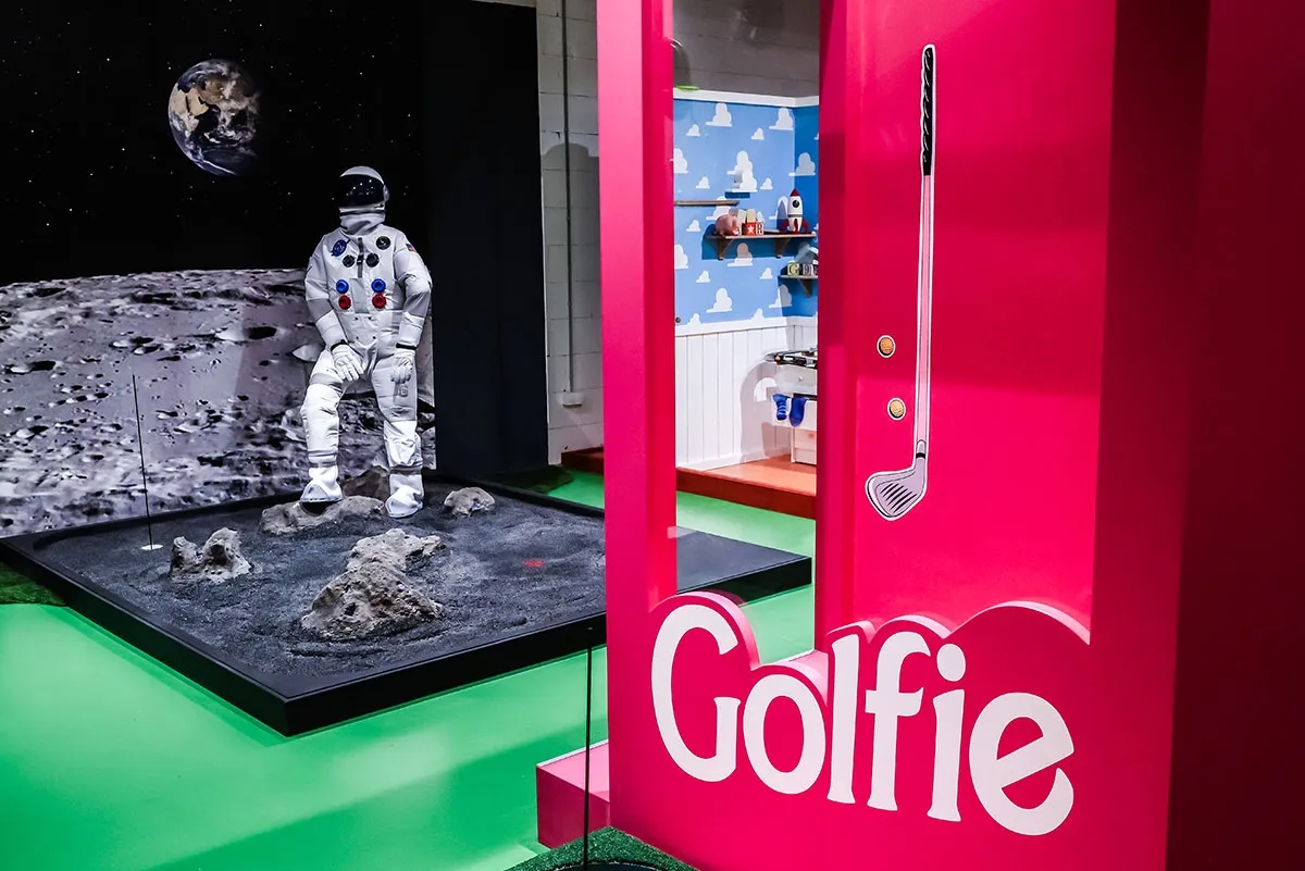 Der Eingangsbereich von Golfzone zeigt eine moderne Leuchtreklame mit dem Schriftzug „Golfie“ und einen durchsichtigen Golfballspender.