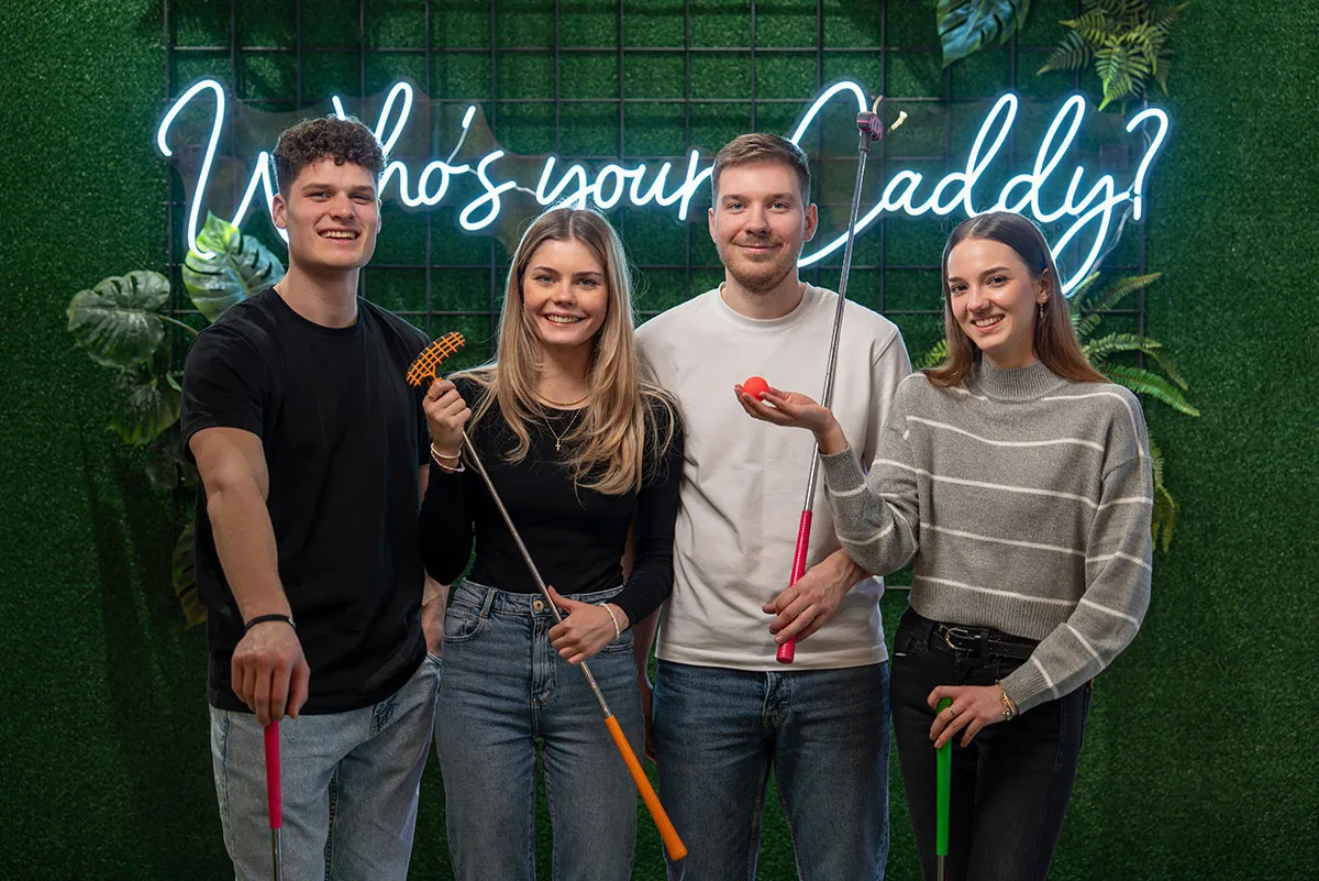 Vier Personen mit Schlaegern vor Neonwand „Who’s your caddy?“