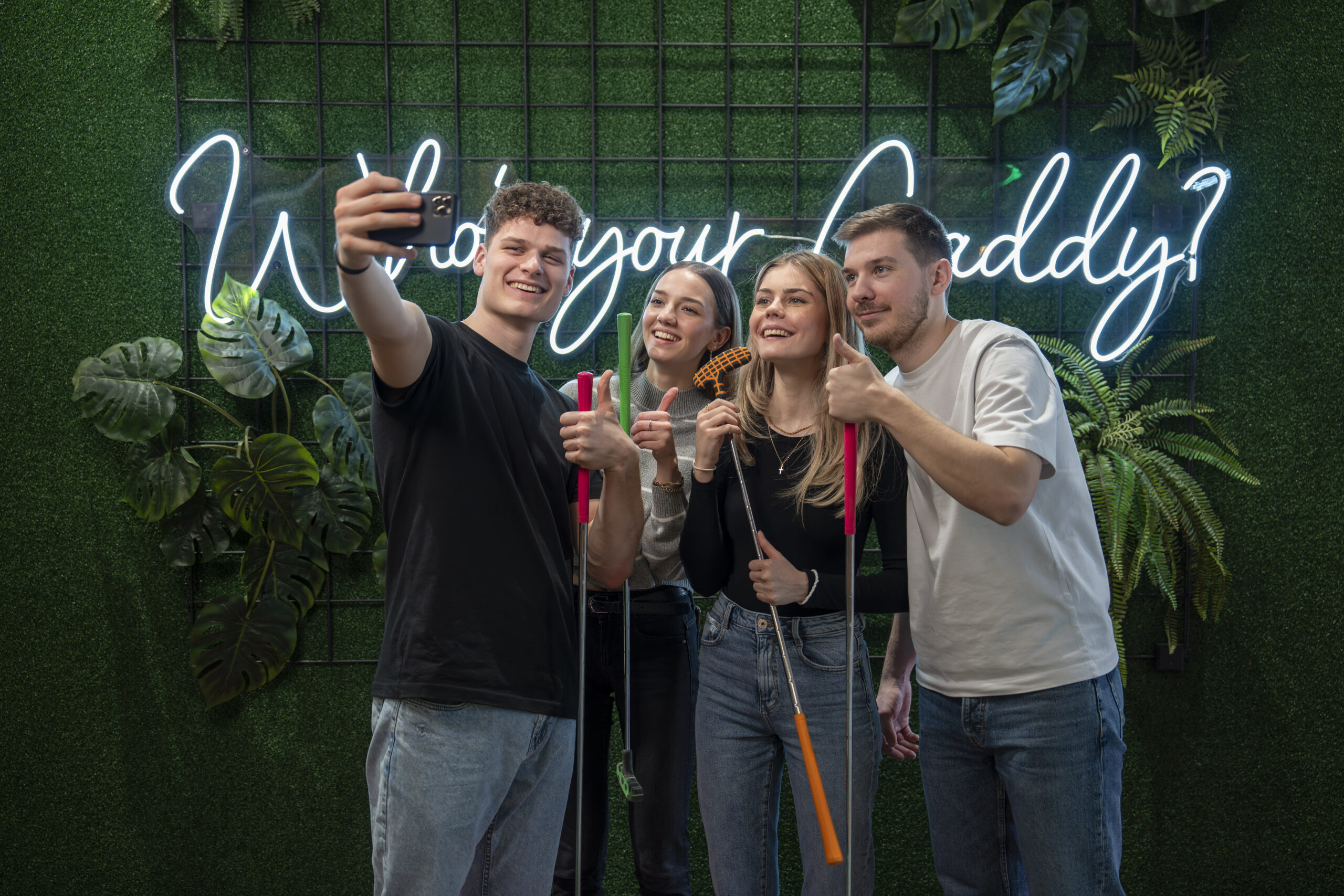 Eine Freundesgruppe, die mit Minigolfschlägern und Minigolfbällen ein Selfie vor einem Neonschild macht.