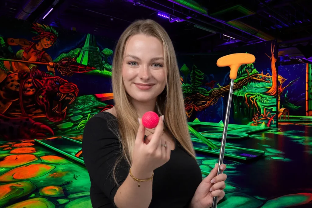 Frau mit pinkem Ball und orangem Schlaeger vor Schwarzlicht Minigolf Kulisse