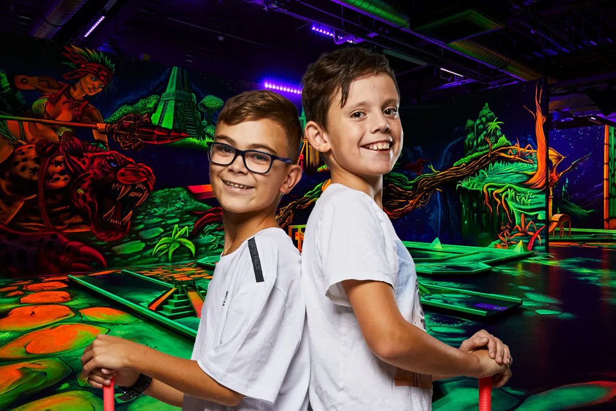 Zwei Jungen stehen freundlich nebeneinander und halten farbige Minigolfschläger. Im Hintergrund sieht man die Schwarzlicht Minigolf Bahn der GolfZone.