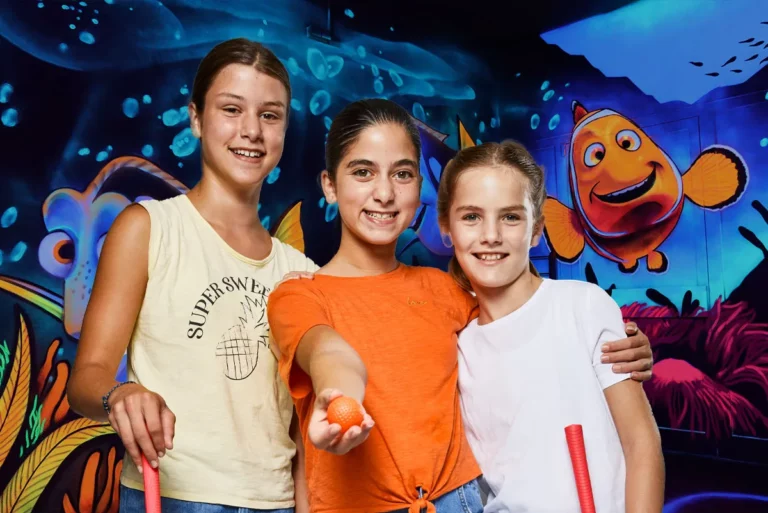 Drei Mädchen posieren fröhlich vor Unterwasser-Wandmotiv im Schwarzlicht-Minigolfbereich