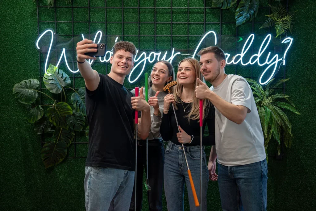 Eine Freundesgruppe, die mit Minigolfschlägern und Minigolfbällen ein Selfie vor einem Neonschild macht.